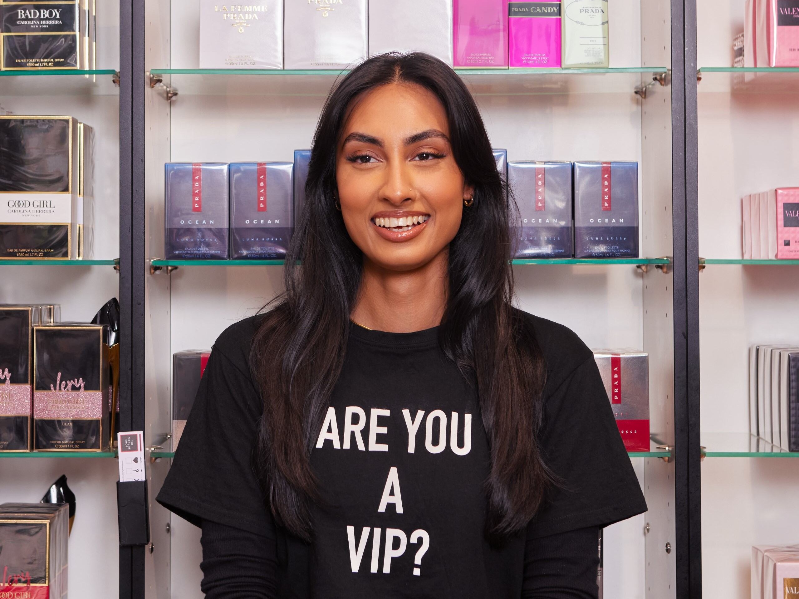 Woman smiling in front of perfume display, wearing a shirt that says "Are you a VIP?"