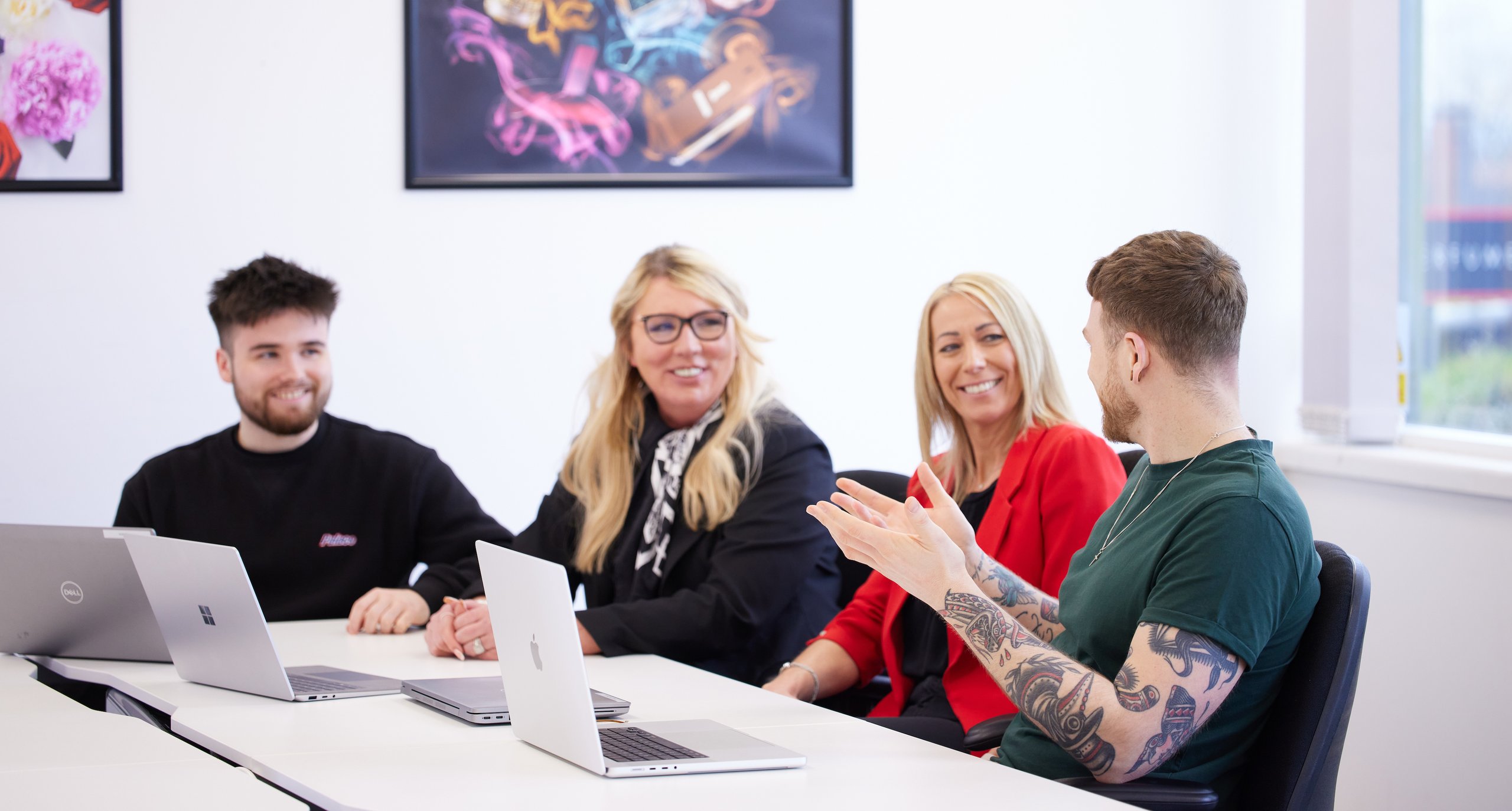 Team members in a meeting at a white table, laptops are present. Wall art decorates the wall.