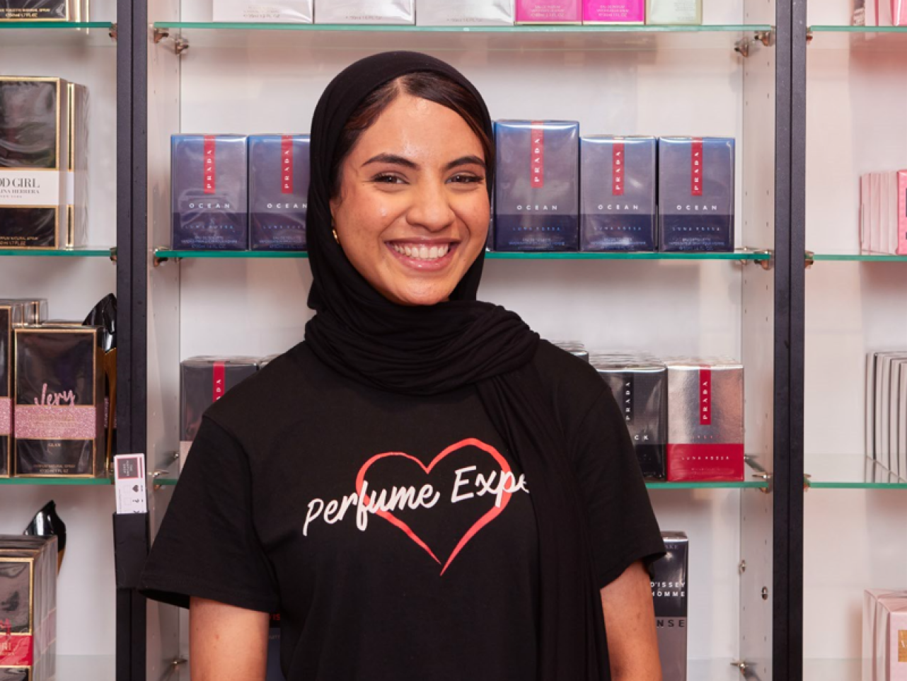 Woman wearing a black hijab and a black "Perfume Expe" shirt smiling in a perfume shop. Shelves with perfume bottles.