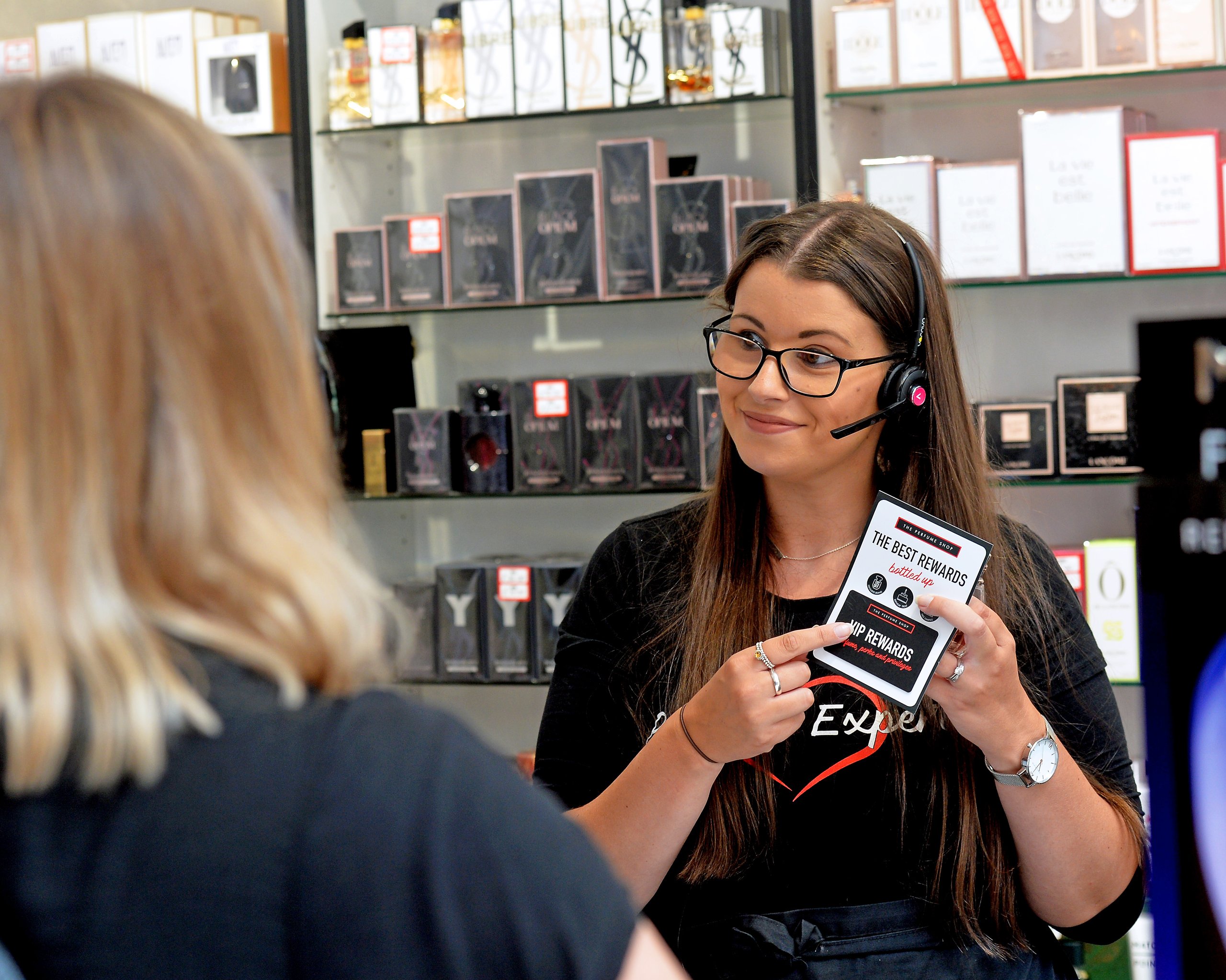 Employee showing a VIP rewards card at The Perfume Shop. Perfume bottles are displayed on shelves.