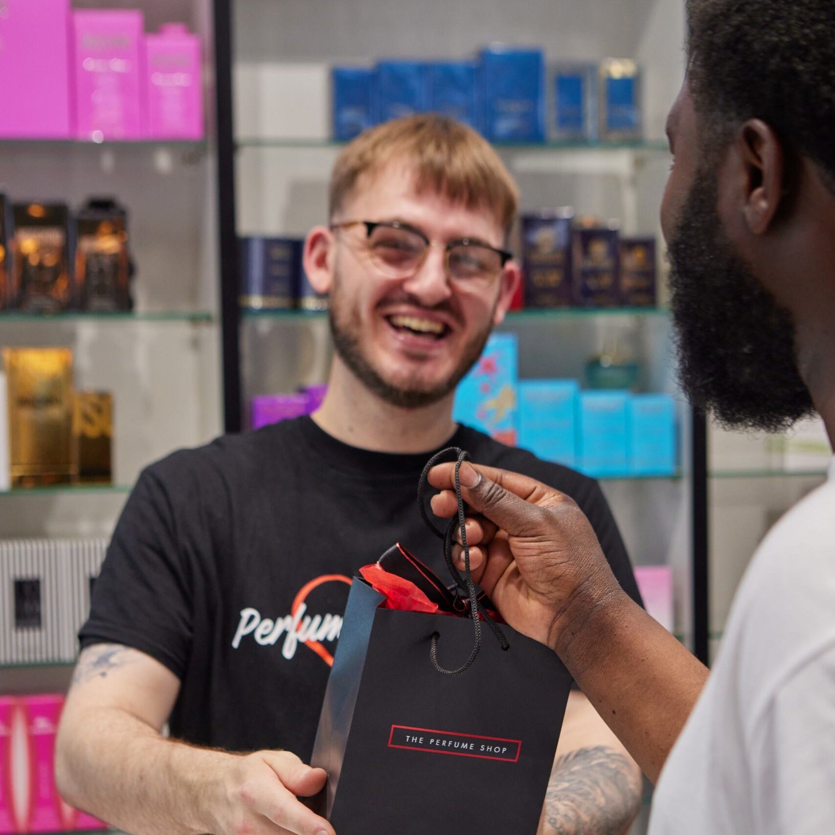 A shop worker hands a black paper bag with red tissue to a customer.