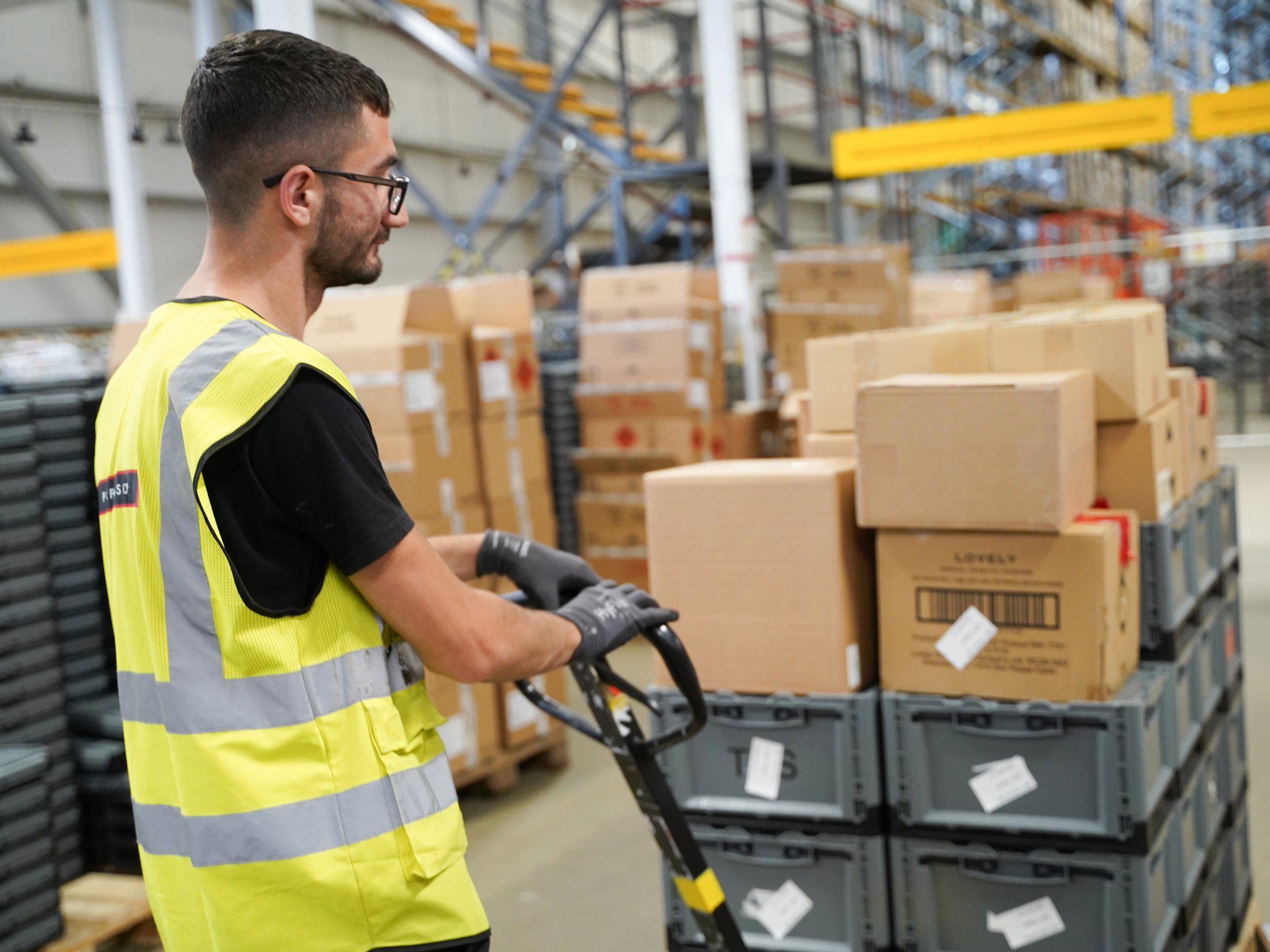 Worker using a pallet jack surrounded by boxes, wearing a safety vest and gloves.