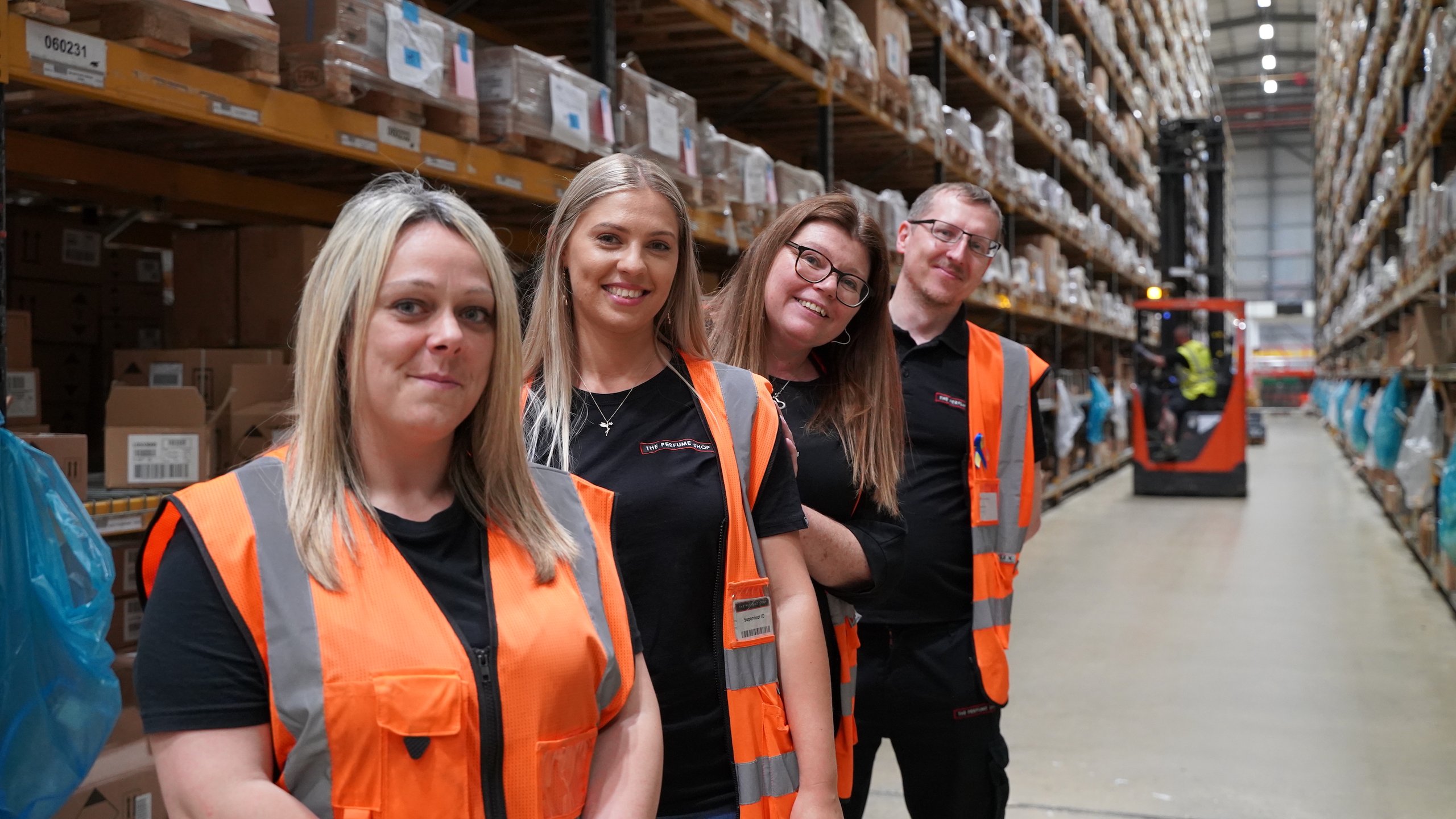 People in high-visibility vests standing in a warehouse. Forklift in background.