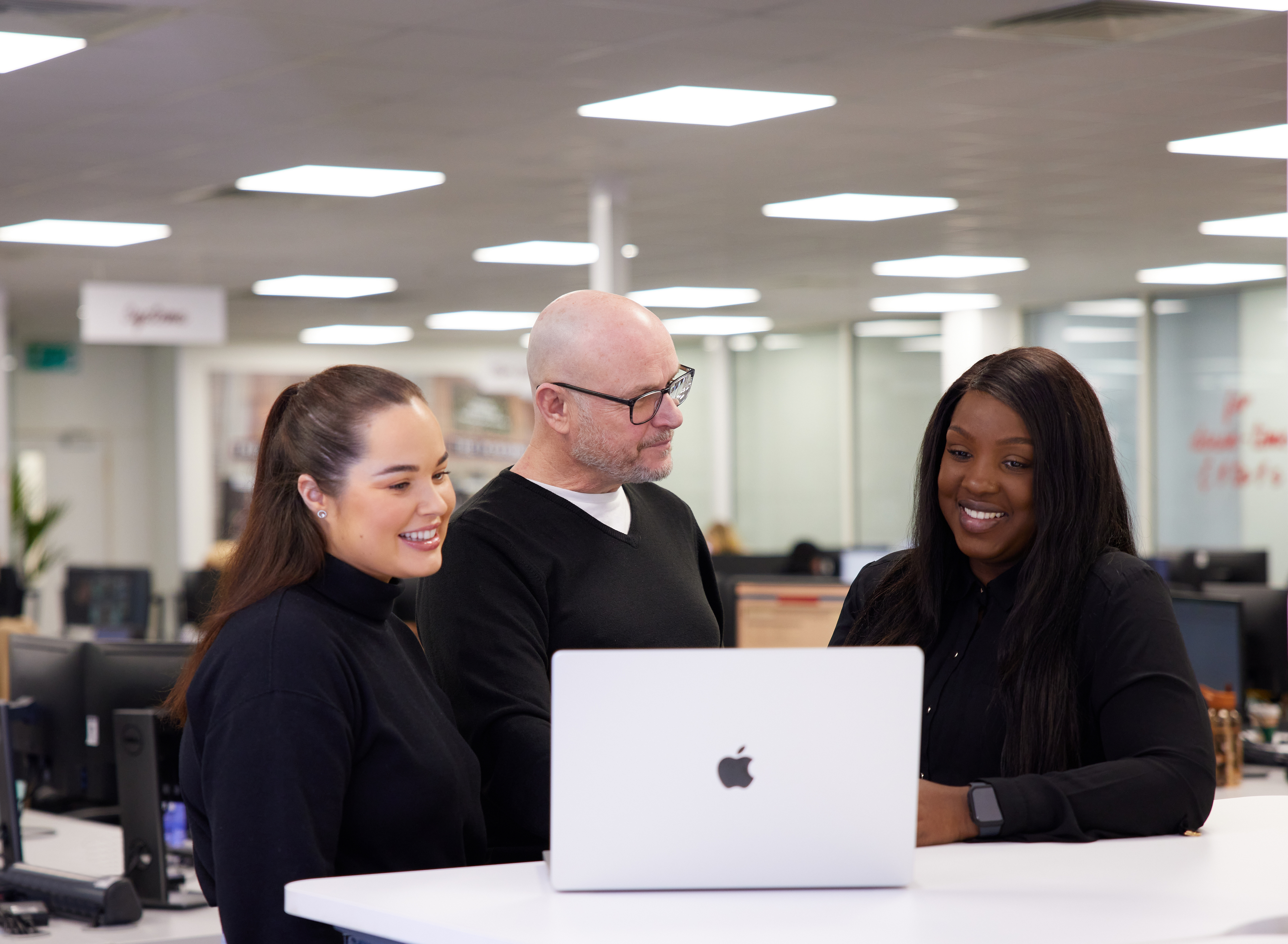 Group of three people looking at a laptop in an office setting.
