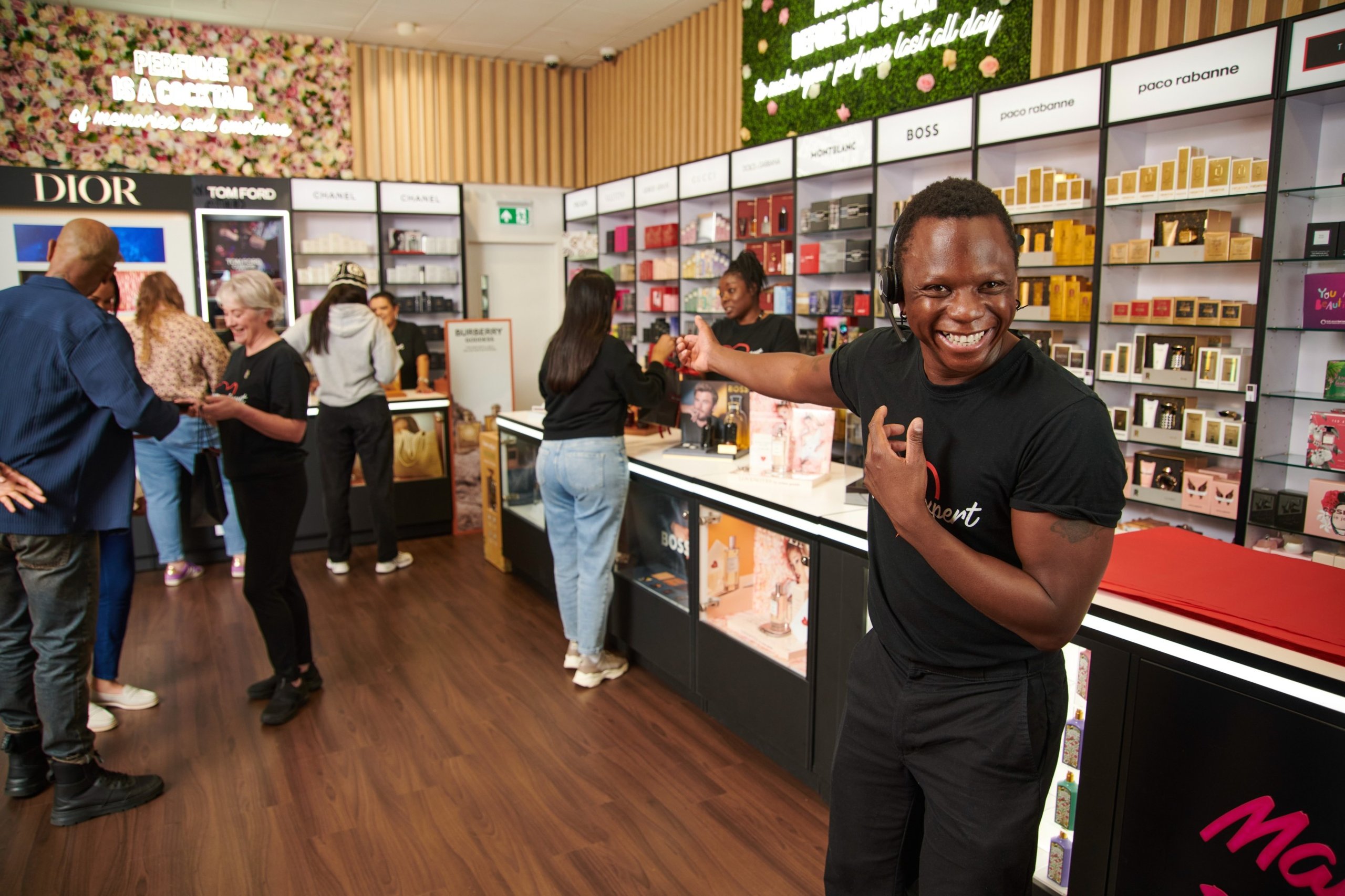 Smiling perfume store employee in front of displays. Customers browse the perfume shop.