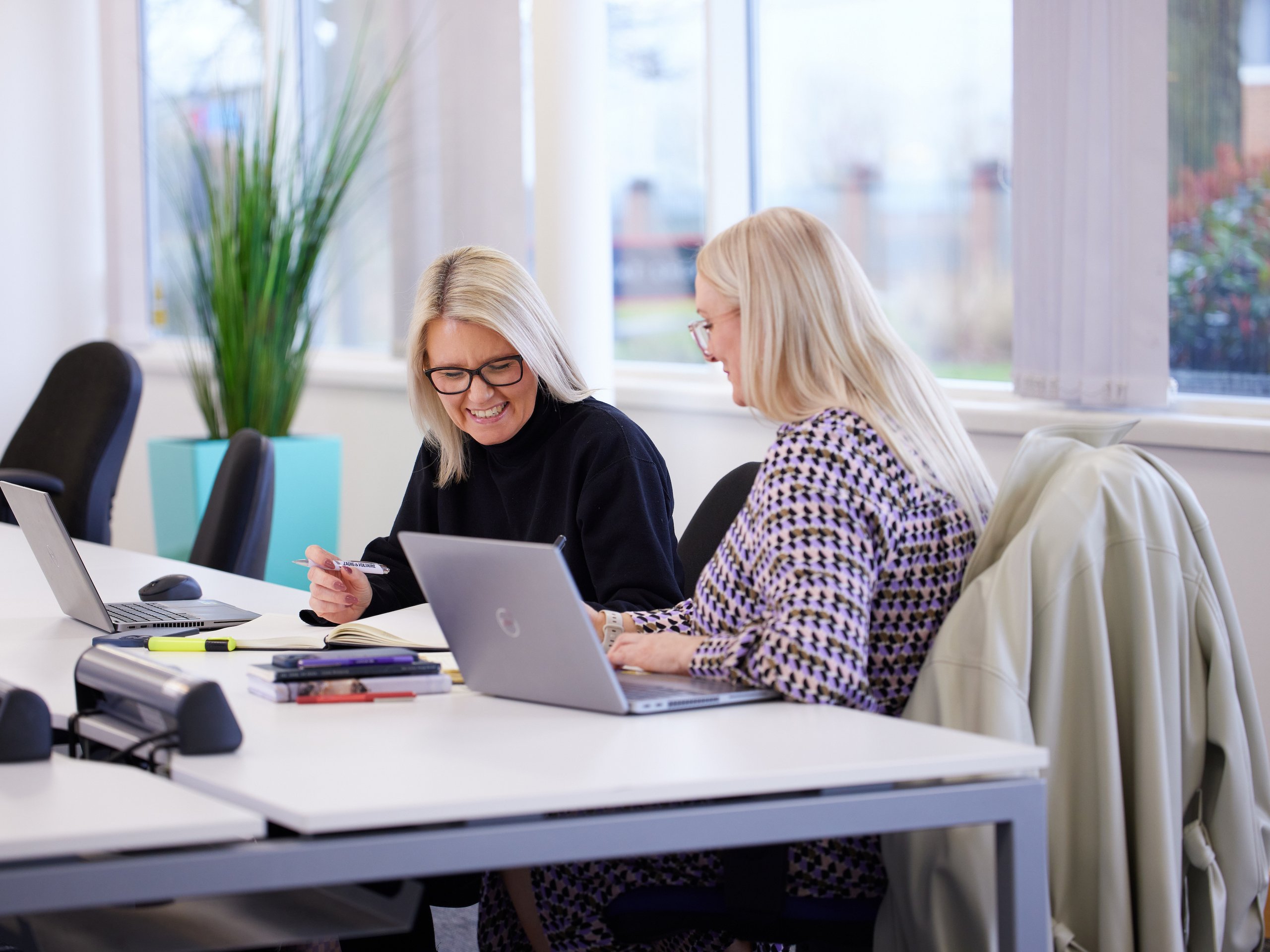 Two women at a desk with laptops, notebook, pen.