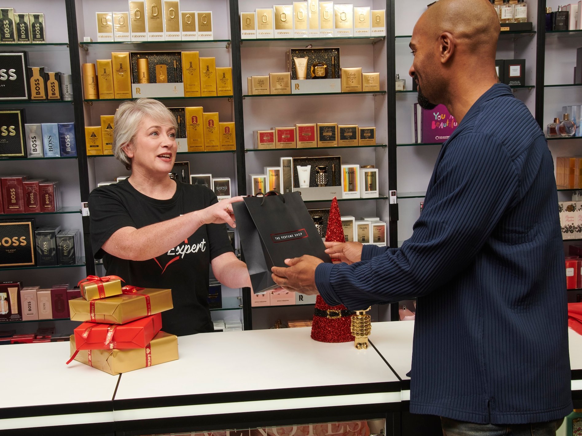 A woman handing a bag to a man in a perfume shop, presents on the counter.