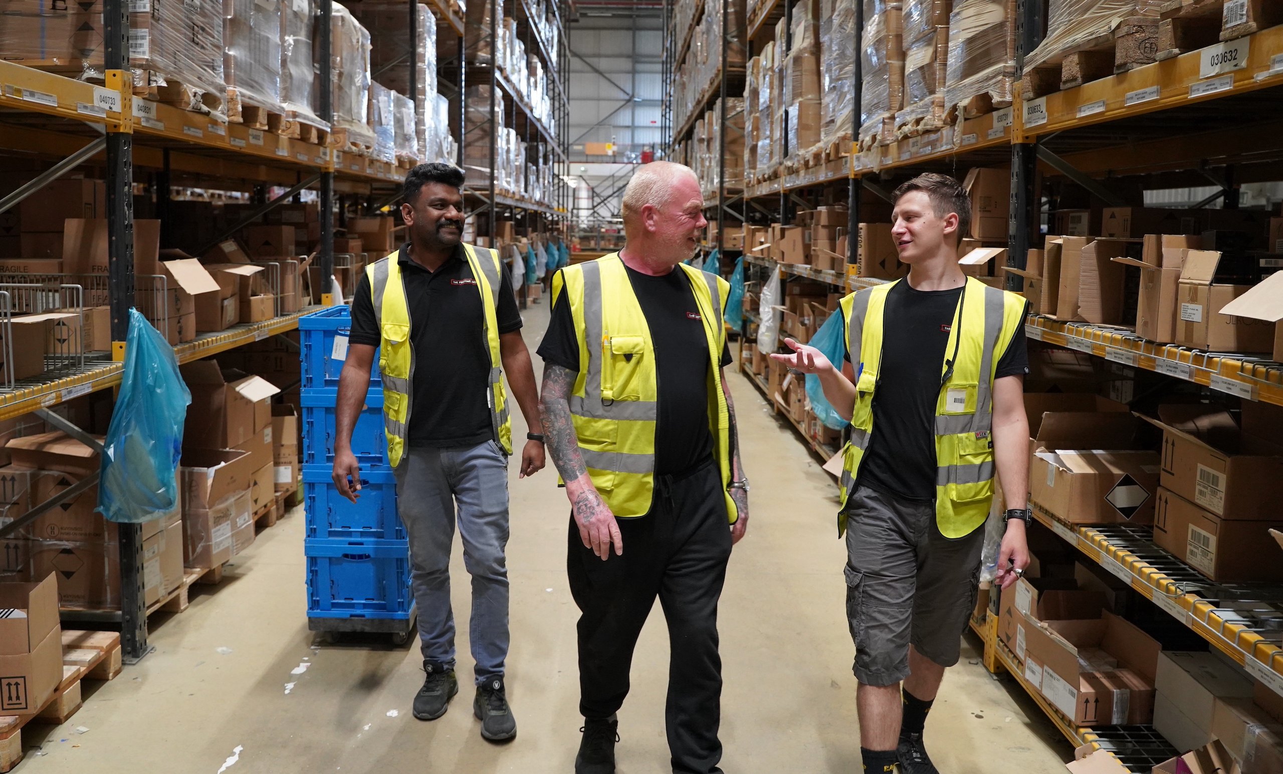 Three men walking in a large warehouse, wearing safety vests. Boxes on shelves.