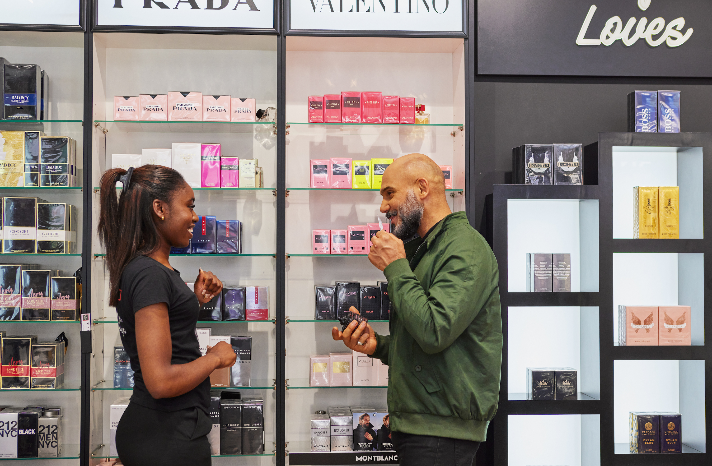 A store employee assists a customer in front of shelves of perfume.