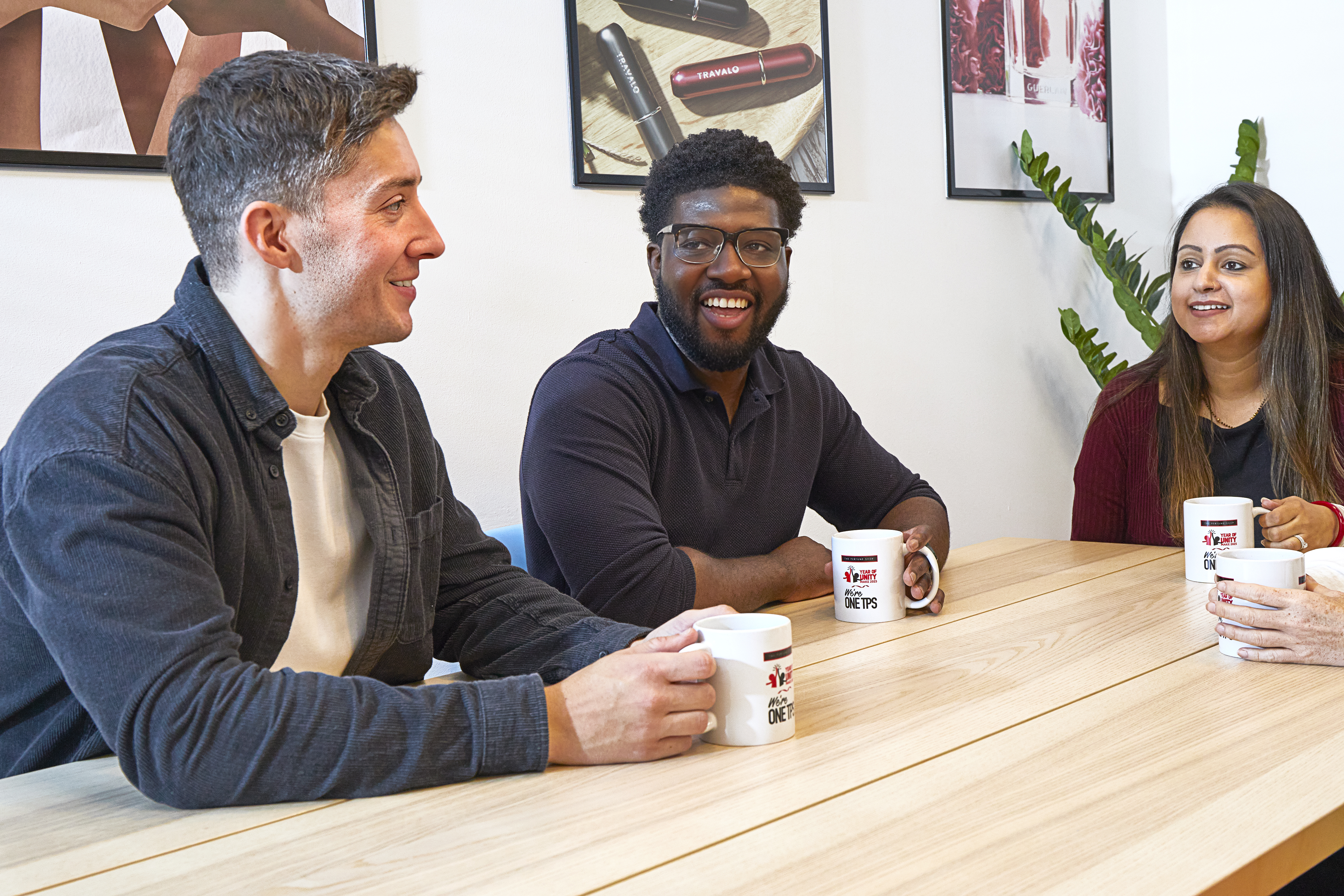 Three colleagues, two men and one woman, are smiling and chatting at a table in an office setting. They are holding mugs with the logo "The Perfume Shop Year of Unity Make 2023 We're ONE TPS."