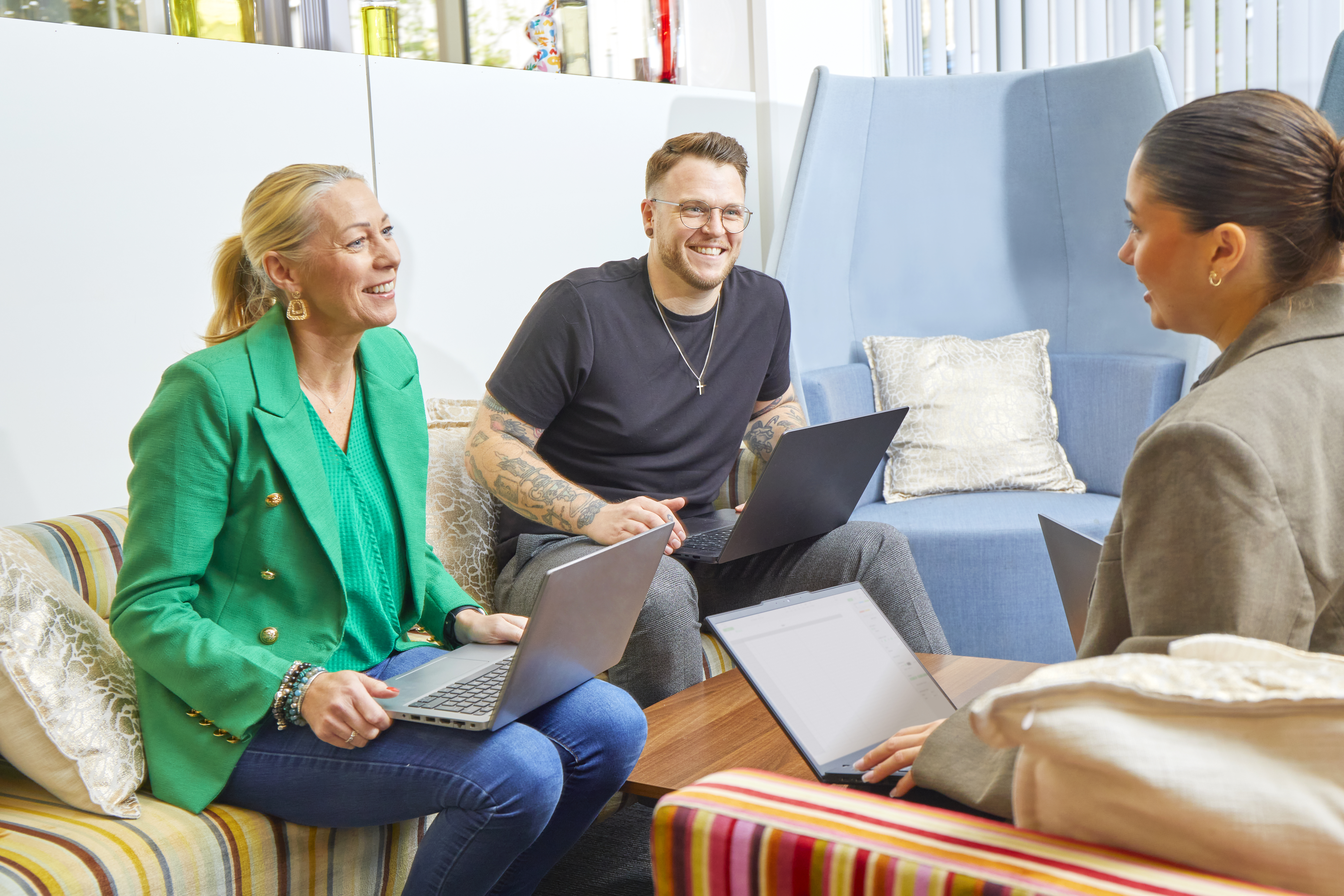 Three colleagues are in a relaxed office setting, working on laptops and discussing ideas.