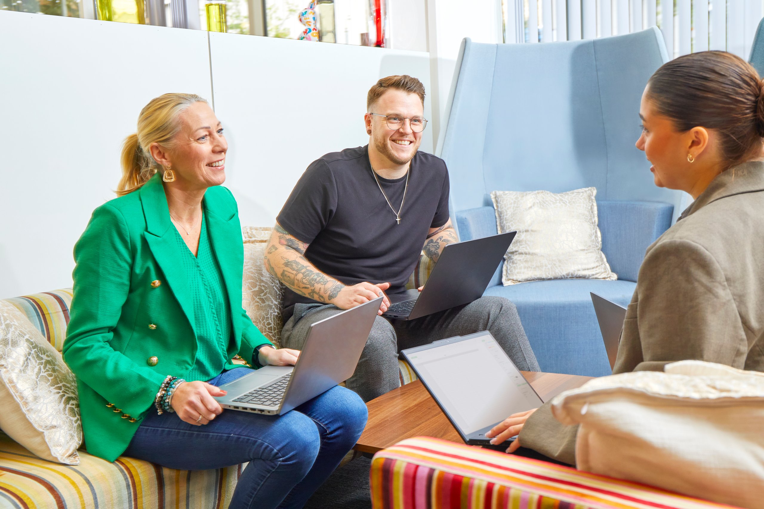 Three colleagues are in a relaxed office setting, working on laptops and discussing ideas.