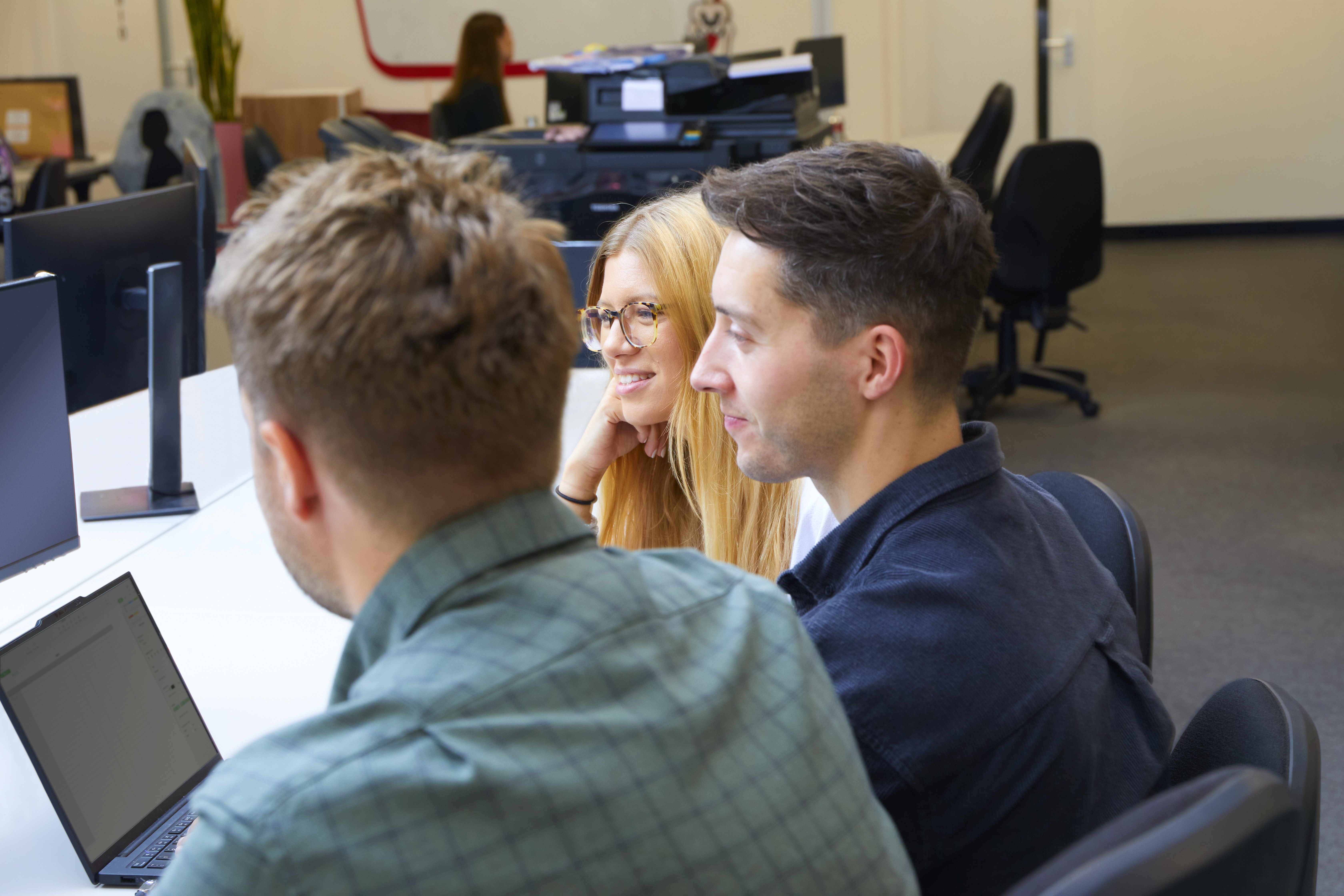 Three colleagues, two men and one woman, are seated at a desk in a modern office, looking at a laptop.